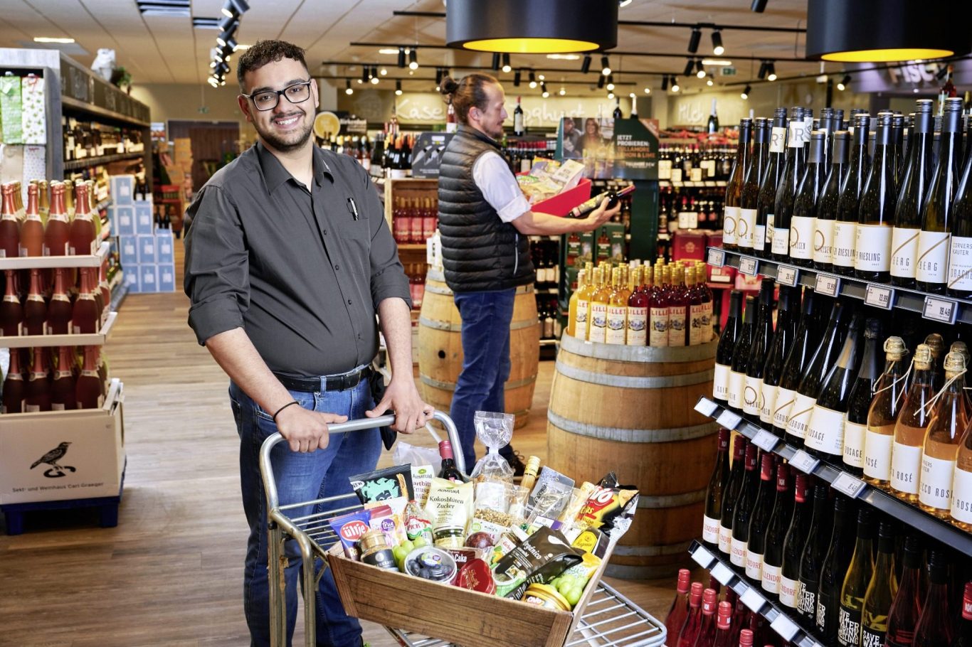 Partyplatten & Geschenkkorbservice Mann mit Einkaufskorb in einem Weingeschäft, Regale mit Wein und Lebensmitteln im Hintergrund.