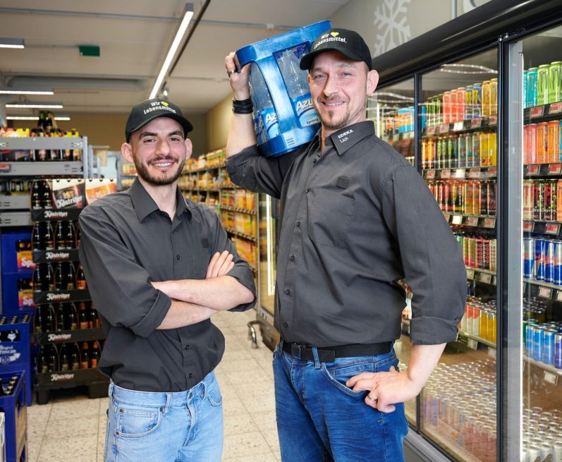 Zwei Männer, einer hält eine Getränkekiste, Regale voller Getränke im Hintergrund.