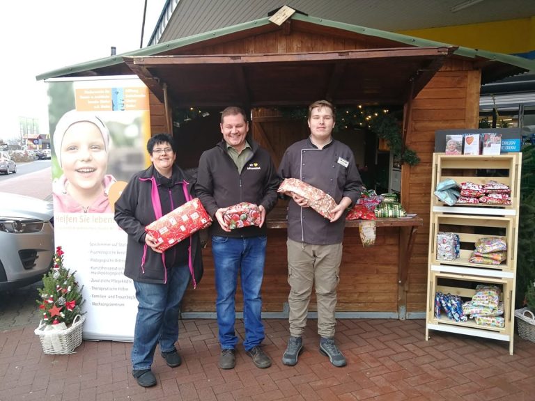 Geschenke für Krebskranke Kinder Mainz e.V. Drei Personen halten Geschenkpakete vor einem Holzhäuschen mit Pflanzen und Informationen.