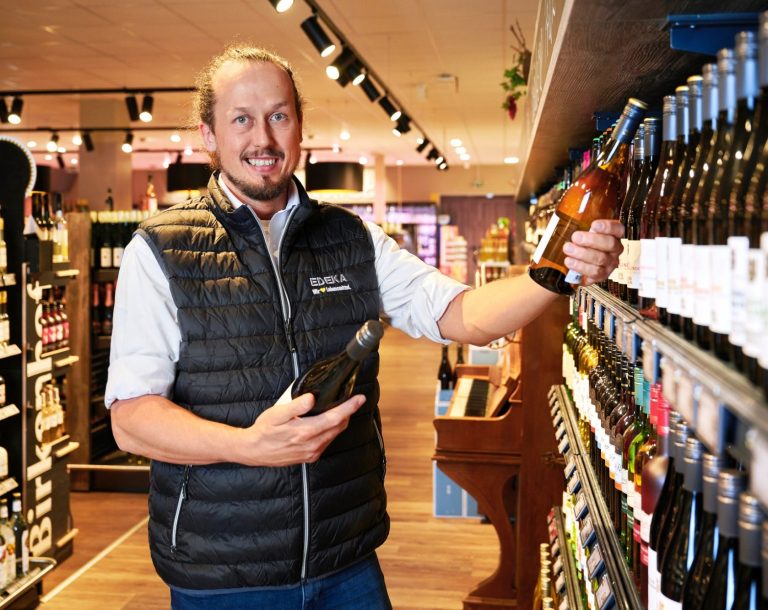 Mann in einem Weingeschäft hält eine Flasche Wein und zeigt auf Regale mit Weinflaschen.