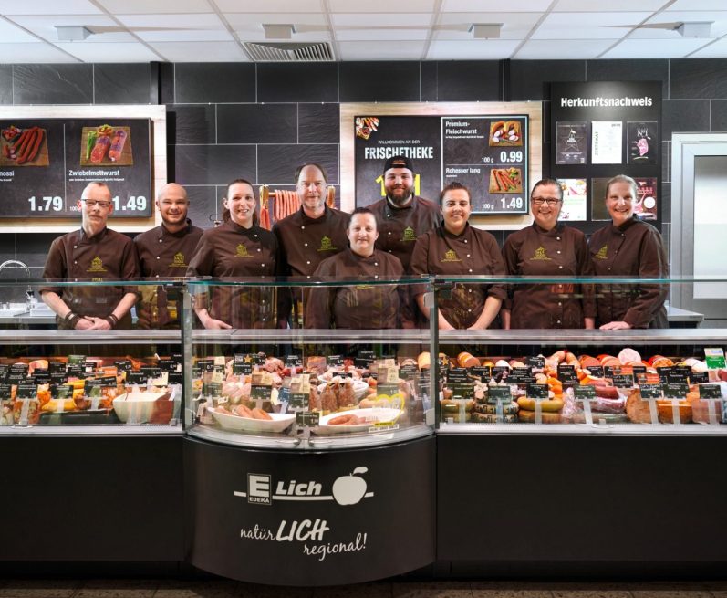 Ein Team von Metzgereimitarbeitern steht vor einer Vitrine mit Wurst und anderen Speisen.